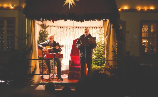 Zwei Männer auf einer Bühne in weihnachtlichem Ambiente und mit einer Gitarre und einem Liederheft © Henry Tornow Zwei Männer auf einer Bühne in weihnachtlichem Ambiente und mit einer Gitarre und einem Liederheft © Henry Tornow