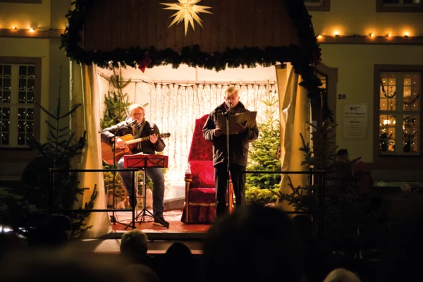 Zwei Männer auf einer Bühne in weihnachtlichem Ambiente und mit einer Gitarre und einem Liederheft