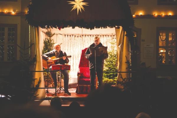 Zwei Männer auf einer Bühne in weihnachtlichem Ambiente und mit einer Gitarre und einem Liederheft © Henry Tornow Zwei Männer auf einer Bühne in weihnachtlichem Ambiente und mit einer Gitarre und einem Liederheft © Henry Tornow